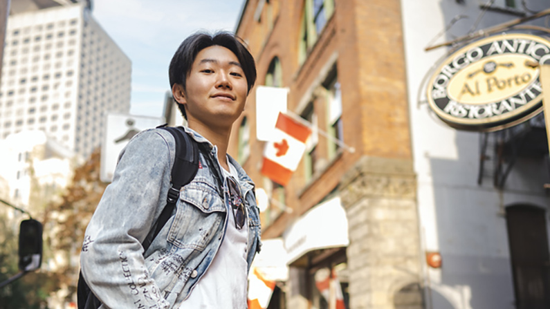 A student stands in downtown location with a Canada flag swaying in the background attached to a building