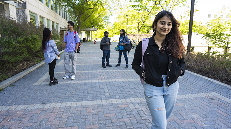 A student stands facing the camera smiling while others behind her chat amongst themselves on campus outdoors