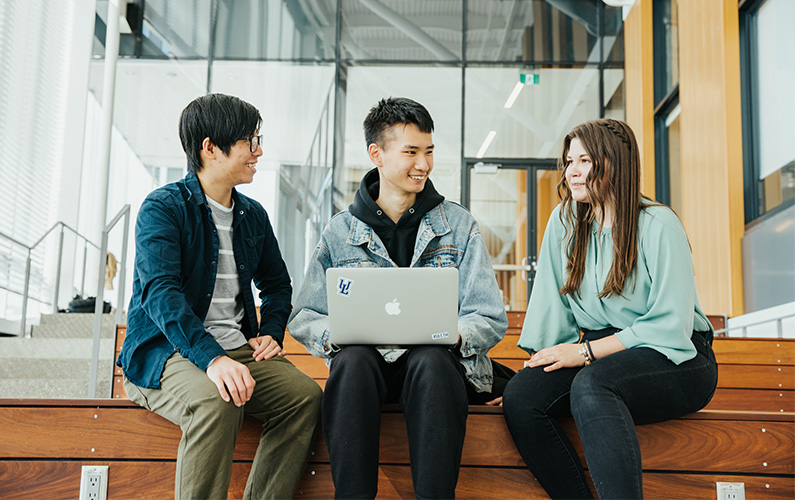 3 students with different ethnicity sitting on a bench and looking at each other. The middle person is holding a laptop.